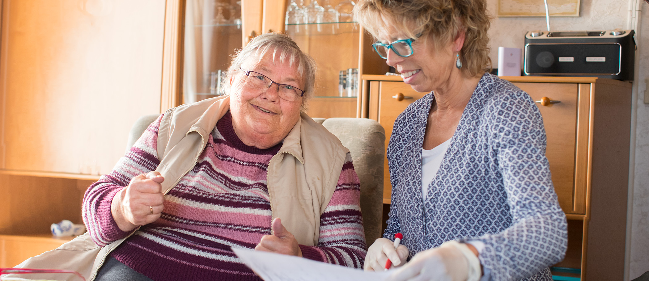 Eine Frau sitzt mit ihrer Pflegerin im Wohnzimmer uns lächelt in die Kamera